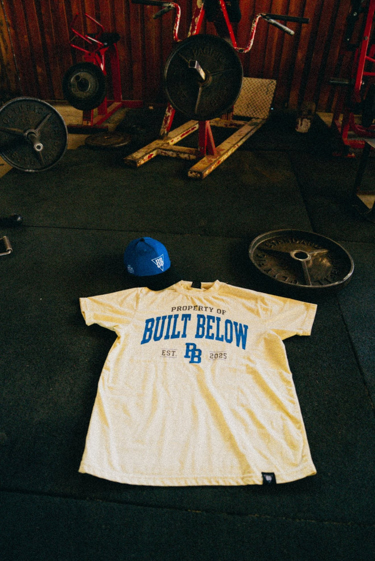 Built Below Varsity Tee and cap flat-lay in the gym
